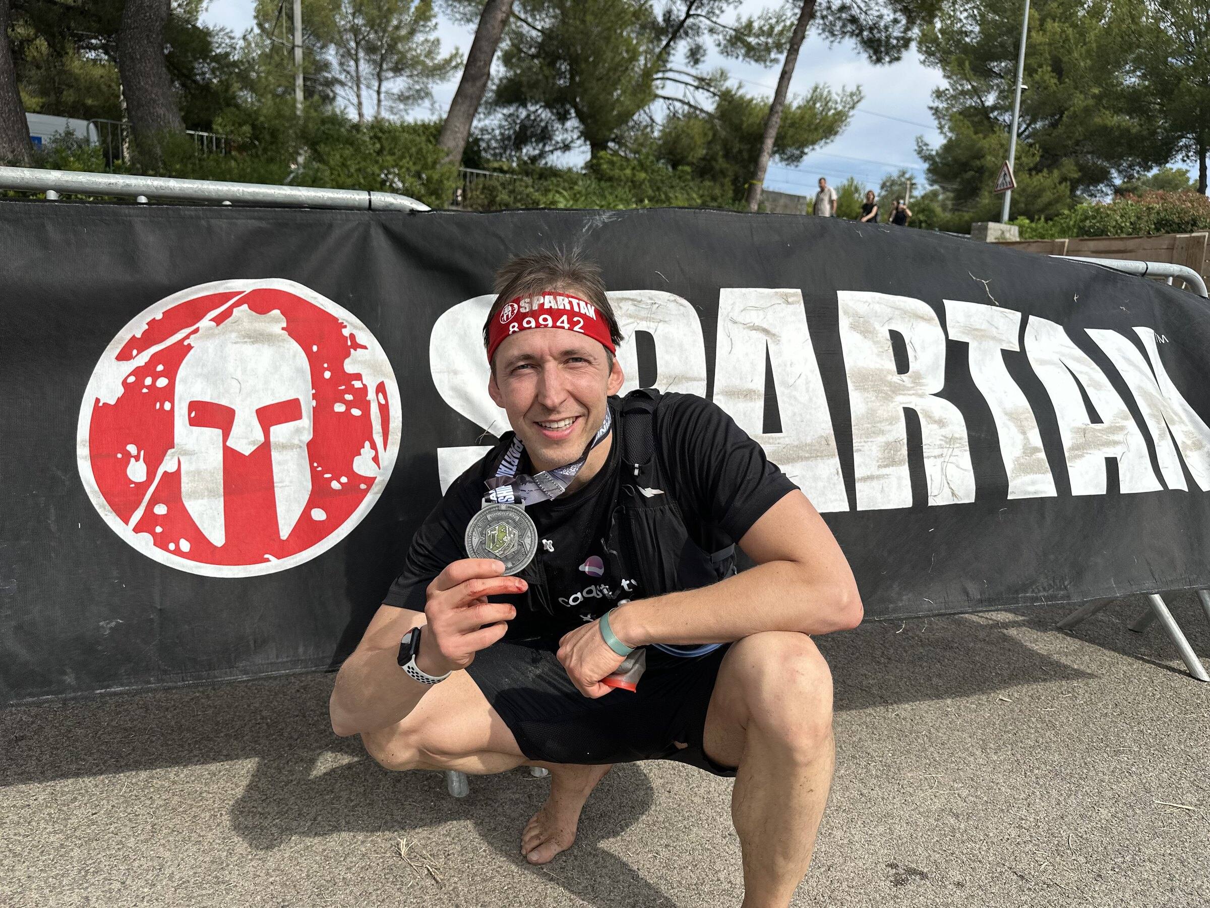 Antoine accroupi pieds nus devant la bâche Spartan, médaille de finisher à la main